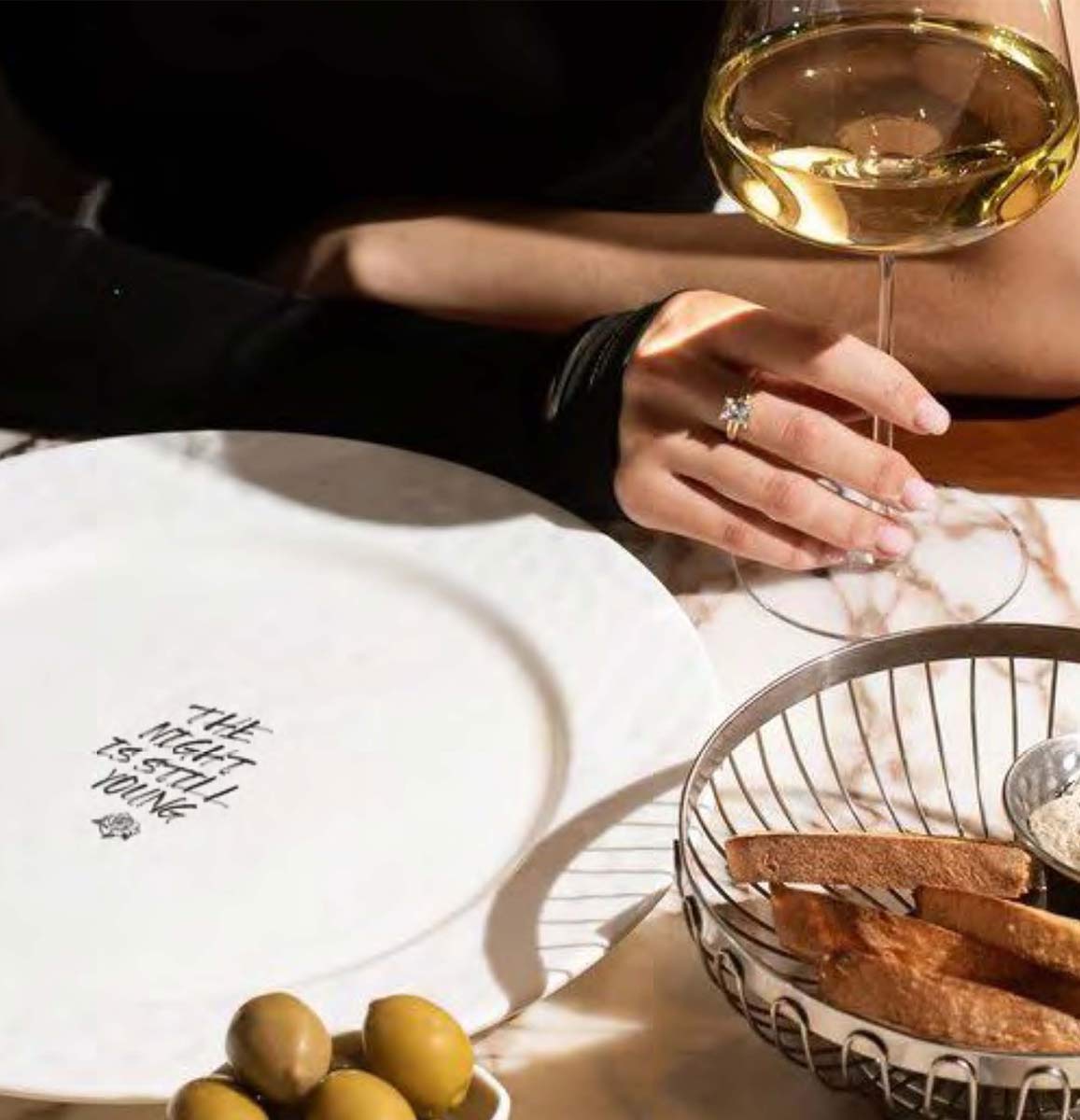 Hand holding stem of glass of white wine with plate on table reading 'The night is still young.'