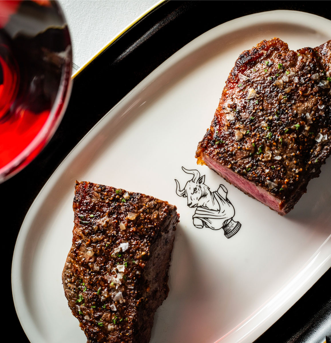 Grilled steak cut in half on presentation plate imprinted with Butcher & Rose bull icon.