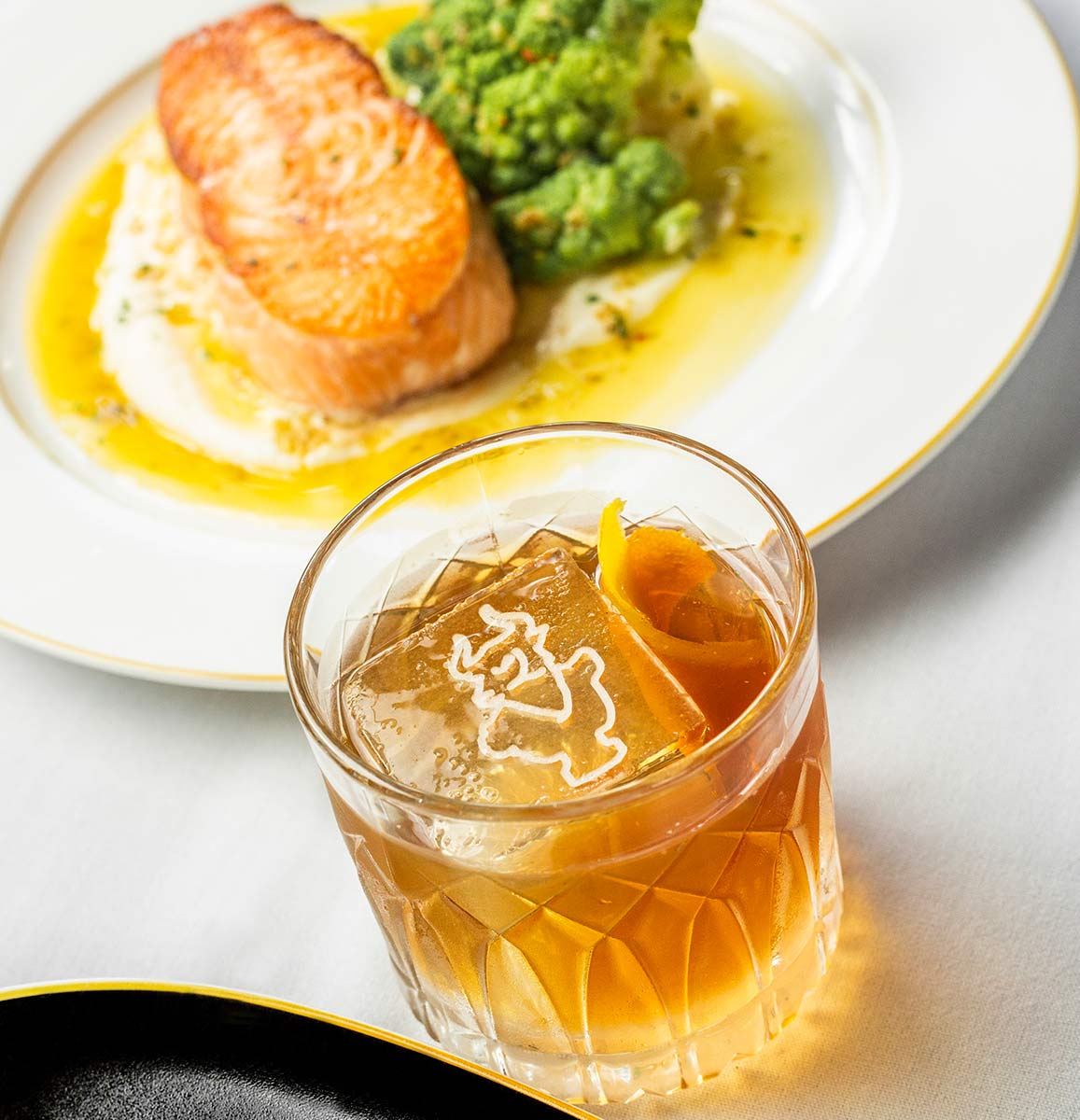 Bourbon cocktail with large square ice cube imprinted with Butcher & Rose logo next to Salmon Steak served at Butcher & Rose in Columbus, Ohio.